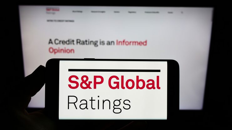 Ein Smartphone zeigt das Logo von S&P Global Ratings mit einem unscharfen Bildschirm im Hintergrund, auf dem der Satz "A Credit Rating is an Informed Opinion" (Ein Kreditrating ist eine fundierte Meinung) zu lesen ist, der zeigt, wie DeFi-Trends die traditionelle Kreditbewertung verändern.
