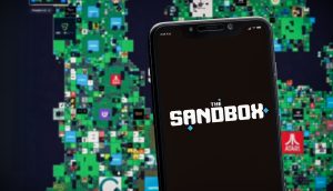 Auf einem Smartphone ist das Logo von The Sandbox zu sehen, im Hintergrund eine digitale Karte der virtuellen Sandbox-Grundstücke mit verschiedenen Logos.