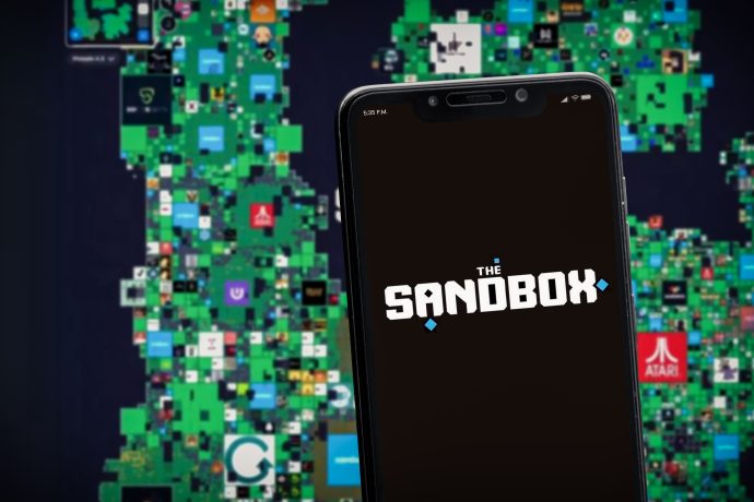 Auf einem Smartphone ist das Logo von The Sandbox zu sehen, im Hintergrund eine digitale Karte der virtuellen Sandbox-Grundstücke mit verschiedenen Logos.