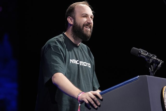 Ein bärtiger Mann spricht auf einem Podium über Bitcoin. Er trägt ein dunkles T-Shirt mit der Aufschrift NAK MOT0 und hat ein Mikrofon vor sich stehen.
