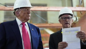 Trump und Powell in Anzügen und weißen Schutzhelmen stehen in einem Haus; einer hält ein Dokument in der Hand und erklärt es, während der andere aufmerksam zuhört, was eine professionelle Geschäftsumgebung in den USA widerspiegelt.
