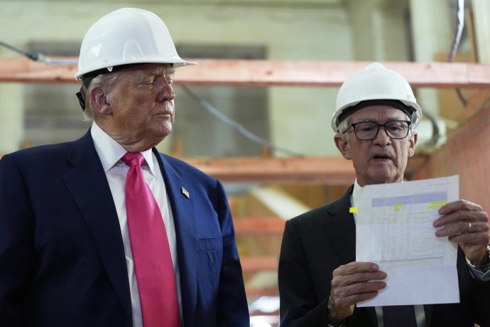 Trump und Powell in Anzügen und weißen Schutzhelmen stehen in einem Haus; einer hält ein Dokument in der Hand und erklärt es, während der andere aufmerksam zuhört, was eine professionelle Geschäftsumgebung in den USA widerspiegelt.