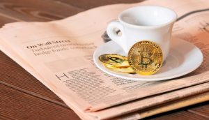 Eine Tasse und eine Untertasse stehen auf einer Zeitung, daneben liegen zwei Krypto-Bitcoin-Münzen auf einer Holzfläche.