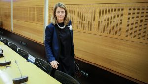 Eine Frau in Geschäftskleidung steht in einem holzgetäfelten Konferenzraum neben einer Reihe von Stühlen und Mikrofonen und bereitet sich auf die Besprechung der neuen MiCA-Vorschriften vor.