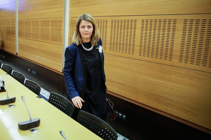 Eine Frau in Geschäftskleidung steht in einem holzgetäfelten Konferenzraum neben einer Reihe von Stühlen und Mikrofonen und bereitet sich auf die Besprechung der neuen MiCA-Vorschriften vor.