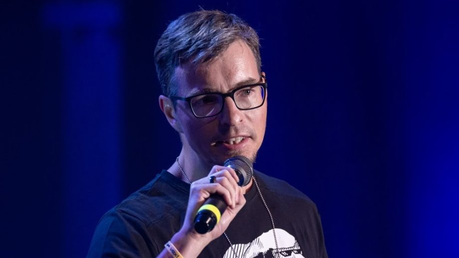 Josef Tětek, die eine Brille und ein schwarzes T-Shirt trägt, spricht auf der Bühne in ein Mikrofon über Bitcoin und hält in der anderen Hand ein Gerät mit blauem Hintergrund.