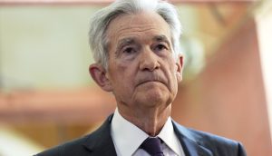 Fed-Chef Powell in Anzug und Krawatte steht drinnen und blickt mit neutraler Miene nach vorne, vielleicht in Erwartung der neuesten Fed-Entscheidung oder der Entwicklungen beim Bitcoin.