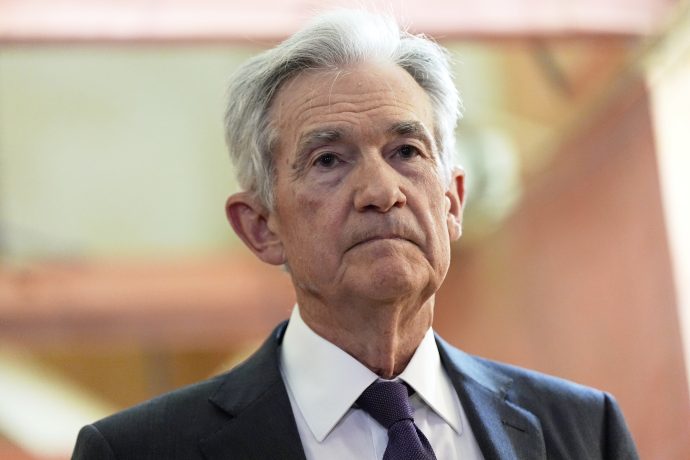 Fed-Chef Powell in Anzug und Krawatte steht drinnen und blickt mit neutraler Miene nach vorne, vielleicht in Erwartung der neuesten Fed-Entscheidung oder der Entwicklungen beim Bitcoin.