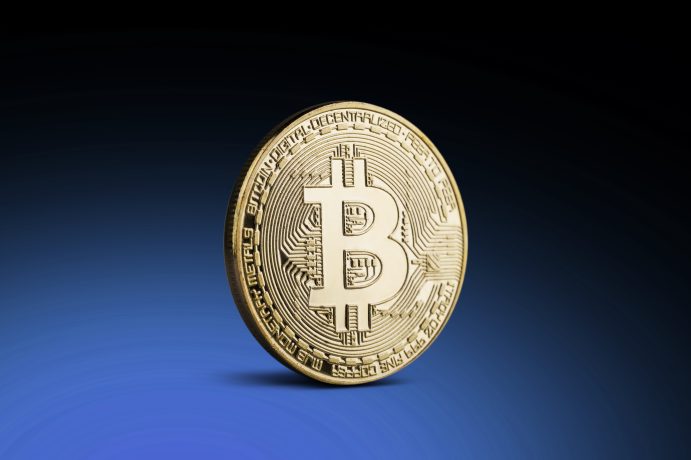 Eine goldfarbene Bitcoin-Münze steht aufrecht auf einer blauen Farbfläche mit kreisförmigen Mustern und Text am Rand, der den digitalen Wert symbolisiert, vielleicht sogar 100.000 Euro.