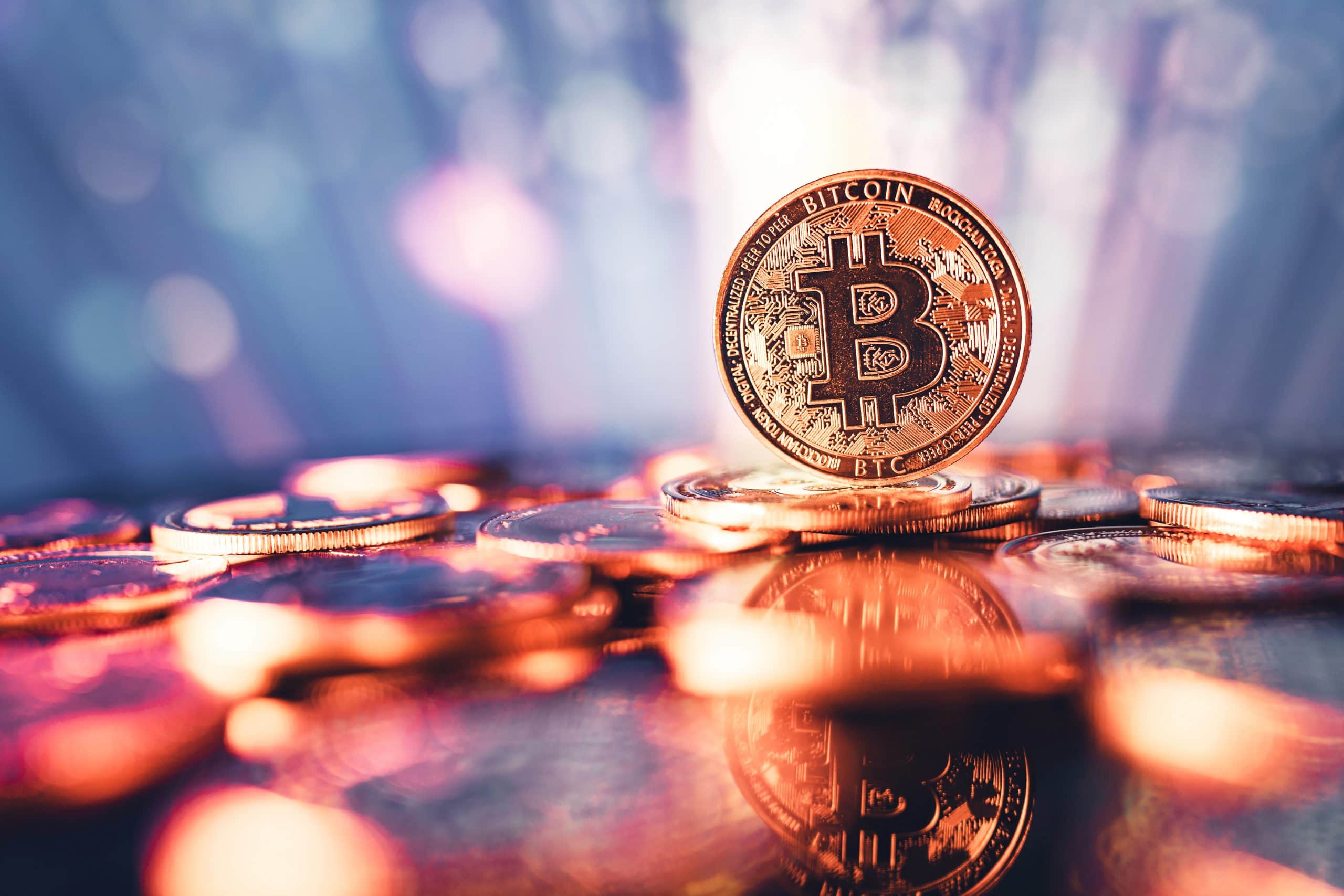 Eine physische Darstellung eines Bitcoins steht aufrecht zwischen verstreuten Münzen, mit einem verschwommenen, bunten Hintergrund und Reflexionen auf der Oberfläche - ein Symbol für die Trends, die in jeder aktuellen Bitcoin-Prognose zu sehen sind.
