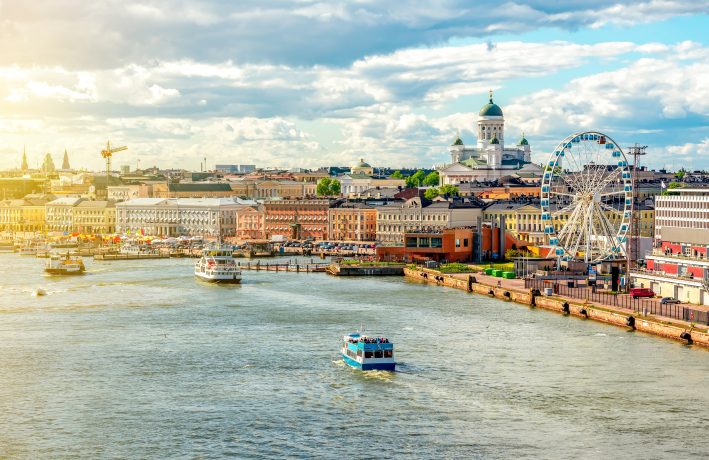 Blick auf die Hafenpromenade von Helsinki mit Booten auf dem Wasser, bunten Gebäuden, einem Riesenrad und dem Dom von Helsinki im Hintergrund bei teilweise bewölktem Himmel - Heimat innovativer Unternehmen wie Tesseract und der lebendigen DeFi-Portfoliomanager-Szene der FIN-FSA.