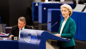 Kommissions-Präsidentin Ursula von der Leyen spricht an einem Podium mit dem Emblem der Europäischen Union über Themen im Zusammenhang mit Russland, während im Hintergrund in einem modernen Konferenzraum ein Mann sitzt und schreibt.