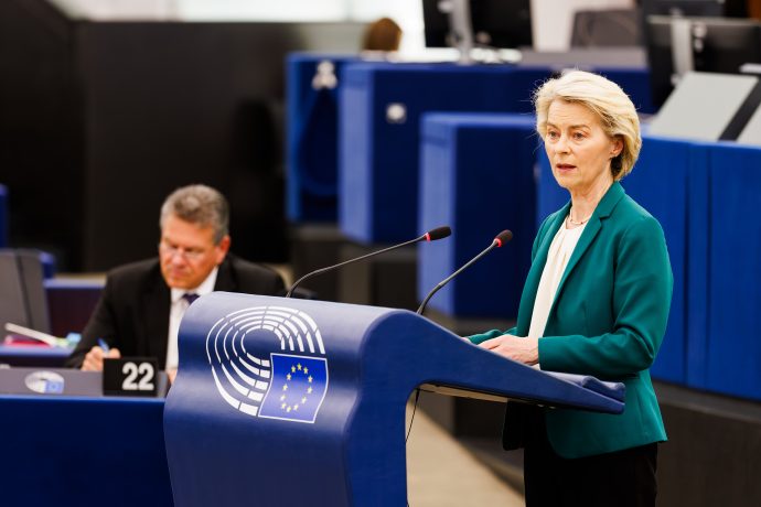 Kommissions-Präsidentin Ursula von der Leyen spricht an einem Podium mit dem Emblem der Europäischen Union über Themen im Zusammenhang mit Russland, während im Hintergrund in einem modernen Konferenzraum ein Mann sitzt und schreibt.