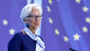 Eine ältere Frau mit weißem Haar, Brille und Kopftuch spricht an einem Podium mit der Aufschrift "EZB" vor einem Hintergrund mit gelben Sternen auf blauem Grund.