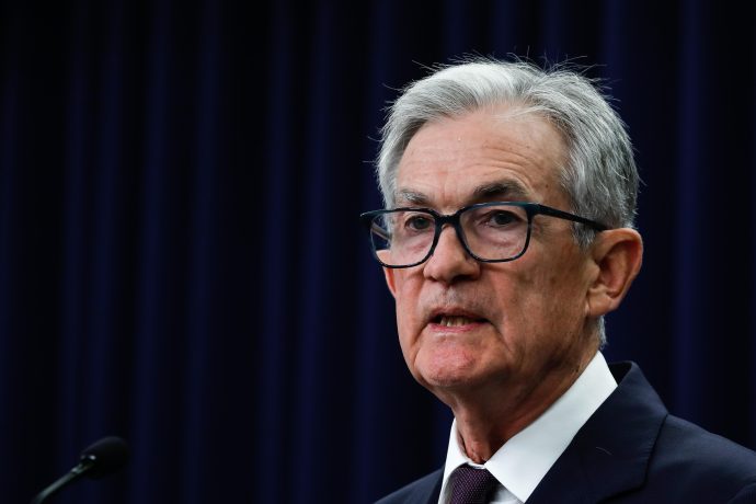 Jerome Powell mit grauem Haar und Brille, der Anzug und Krawatte trägt, spricht an einem Podium vor einem dunklen Vorhang im Hintergrund.