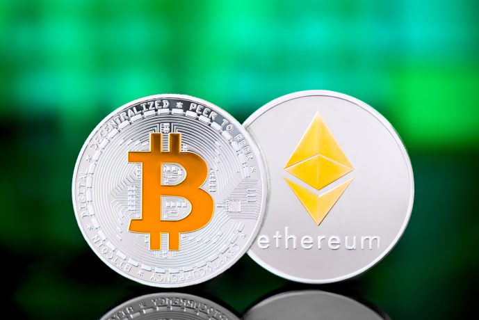 Zwei physische Münzen, darunter eine, die Ethereum repräsentiert, werden nebeneinander vor einem verschwommenen grünen und blauen Hintergrund angezeigt.
