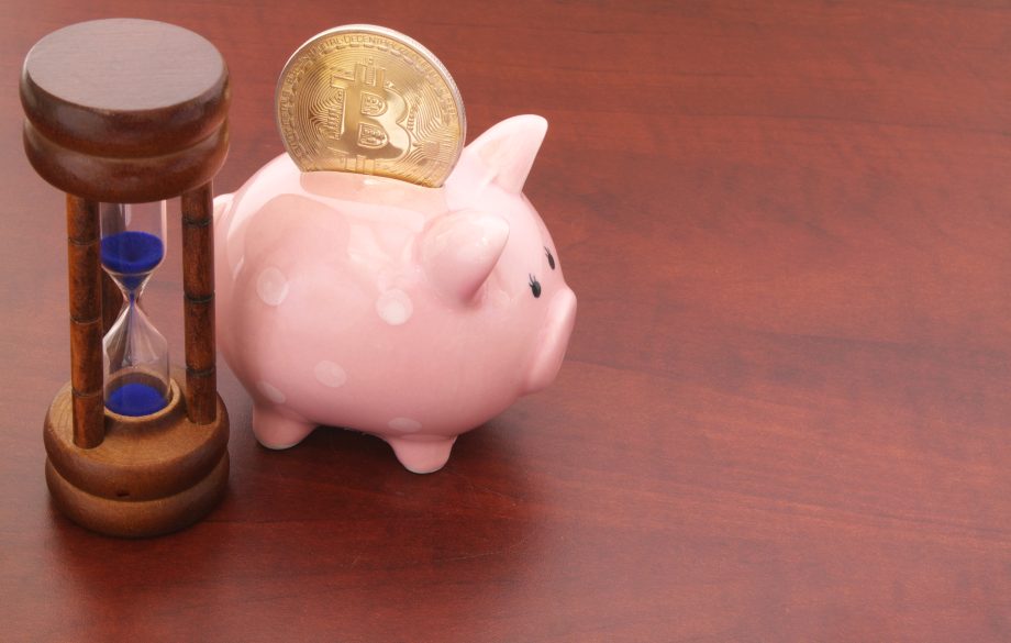 Ein Sparschwein mit einer Bitcoin-Münze darin sitzt neben einer kleinen Sanduhr auf einer hölzernen Oberfläche und symbolisiert das Konzept der Bitcoin-Rente und des Sparens für die Zukunft.