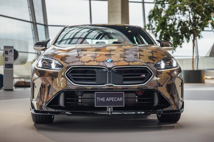 Ein BMW mit einer Schlangenhautverkleidung in Tarnfarben ist im Innenraum ausgestellt und auf einer Frontplatte mit THE APECAR beschriftet - ein Blickfang für jeden Auto- oder NFT-Liebhaber.