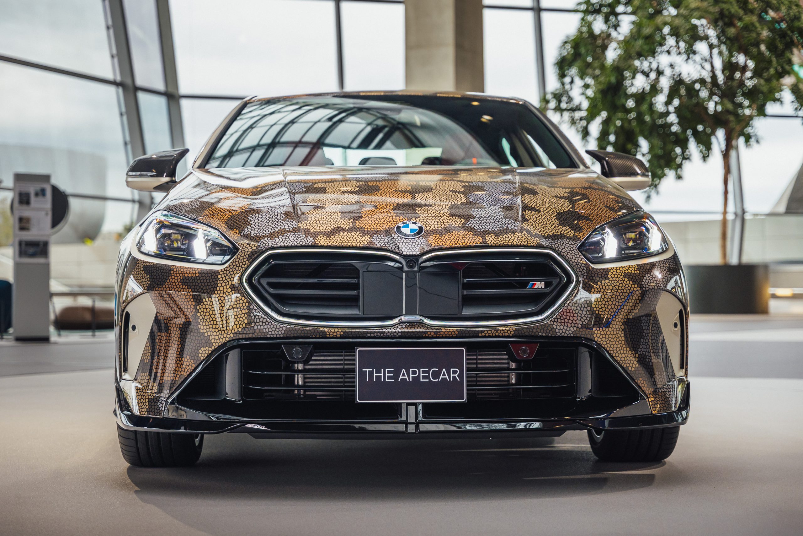 Ein BMW mit einer Schlangenhautverkleidung in Tarnfarben ist im Innenraum ausgestellt und auf einer Frontplatte mit THE APECAR beschriftet - ein Blickfang für jeden Auto- oder NFT-Liebhaber.