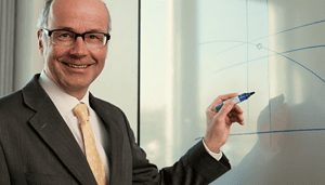 Ein Finanzexperte im Anzug und mit Brille steht an einem Whiteboard, hält einen Marker und zeichnet Bitcoin-Diagramme, während er in die Kamera lächelt.