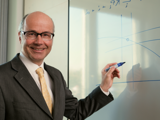 Ein Finanzexperte im Anzug und mit Brille steht an einem Whiteboard, hält einen Marker und zeichnet Bitcoin-Diagramme, während er in die Kamera lächelt.