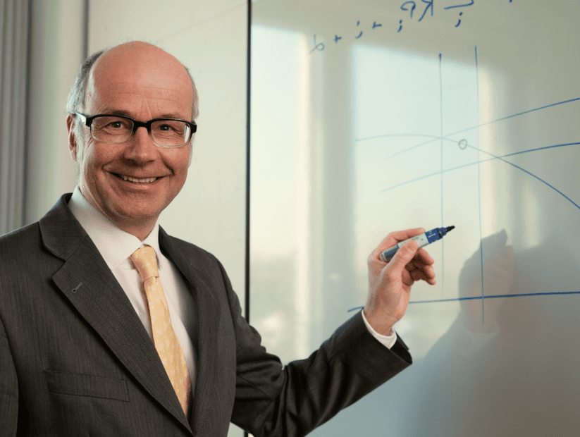 Ein Finanzexperte im Anzug und mit Brille steht an einem Whiteboard, hält einen Marker und zeichnet Bitcoin-Diagramme, während er in die Kamera lächelt.