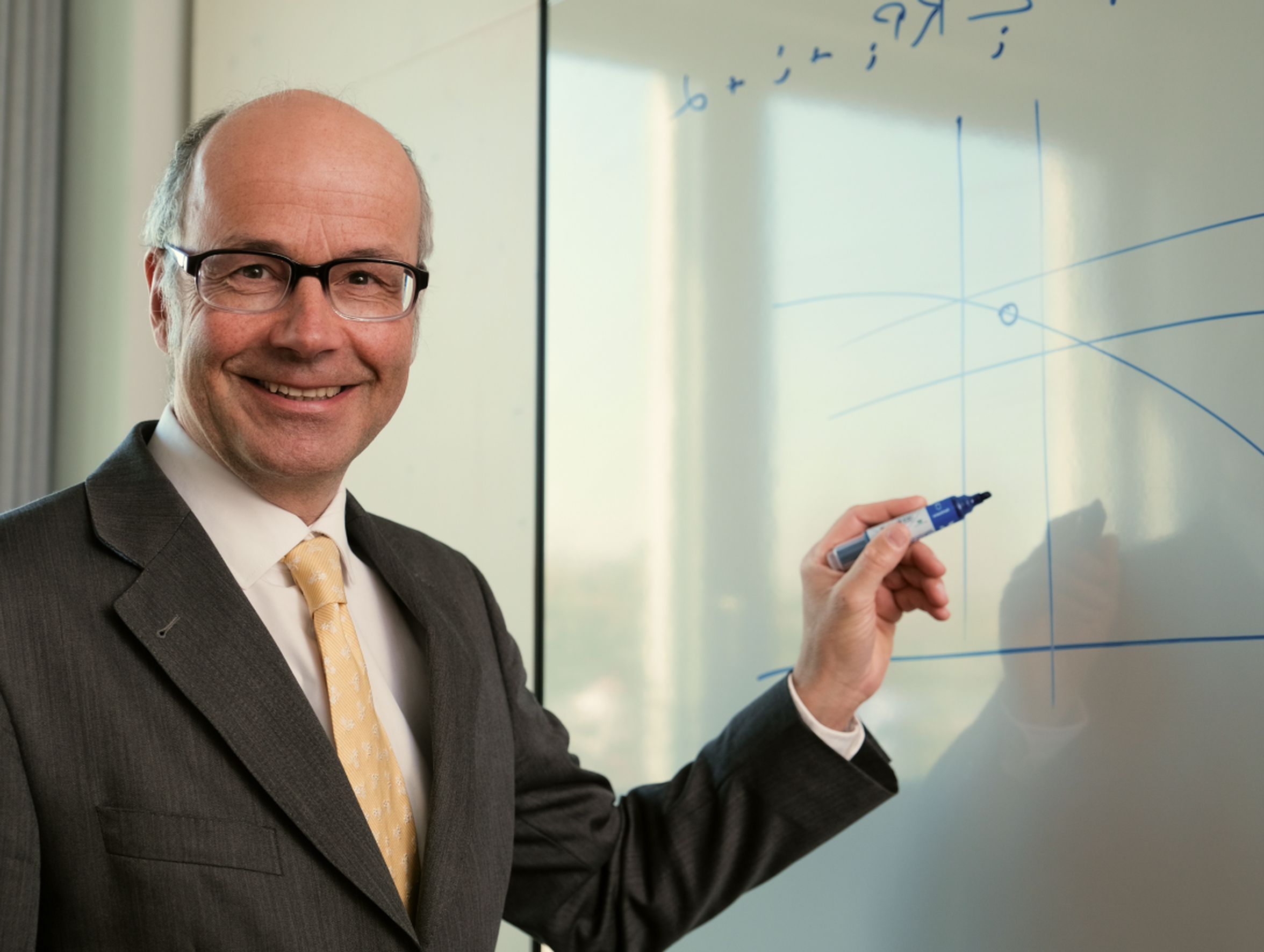 Ein Finanzexperte im Anzug und mit Brille steht an einem Whiteboard, hält einen Marker und zeichnet Bitcoin-Diagramme, während er in die Kamera lächelt.