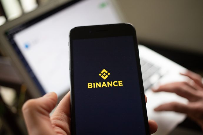 Eine Person hält ein Smartphone mit dem Binance-Logo in der Hand, während im Hintergrund eine Laptop-Tastatur und ein Bildschirm zu sehen sind, was das Interesse an Krypto-Plattformen wie Aster unterstreicht.