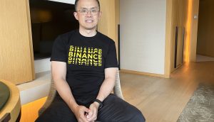 Ein Mann mit Brille und schwarzem Binance-T-Shirt sitzt auf einem Stuhl in einem modernen, gut beleuchteten Raum mit Holzboden und neutralen Wänden und diskutiert darüber, wie nordkoreanische Hacker die Kryptosicherheit im Jahr 2025 beeinflussen könnten.