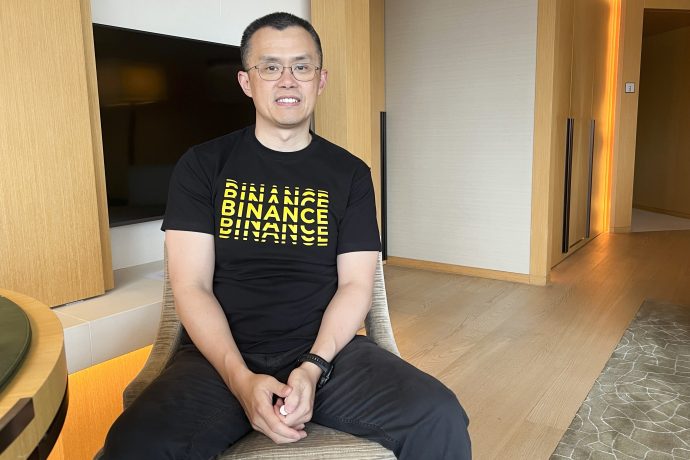 Ein Mann mit Brille und schwarzem Binance-T-Shirt sitzt auf einem Stuhl in einem modernen, gut beleuchteten Raum mit Holzboden und neutralen Wänden und diskutiert darüber, wie nordkoreanische Hacker die Kryptosicherheit im Jahr 2025 beeinflussen könnten.