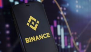 Ein Smartphone mit dem Binance-Logo wird vor einem Bildschirm mit Krypto-Kurs-Charts und Candlestick-Grafiken gezeigt, die neue Rekorde erreichen.