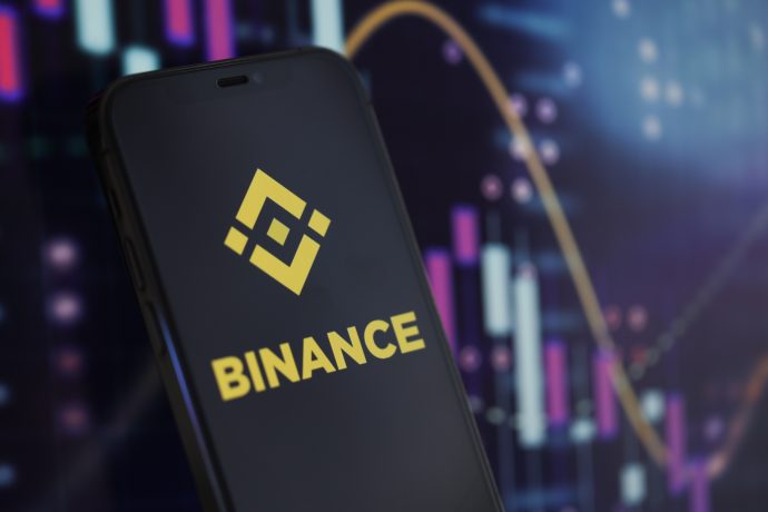 Ein Smartphone mit dem Binance-Logo wird vor einem Bildschirm mit Krypto-Kurs-Charts und Candlestick-Grafiken gezeigt, die neue Rekorde erreichen.