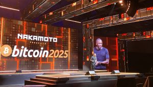 Ein Redner steht auf der Bühne einer Bitcoin 2025-Veranstaltung, während im Hintergrund auf großen Bildschirmen NAKAMOTO PRESENTS bitcoin 2025 zu sehen ist - Arthur Hayes und andere führende Bitcoin-Experten werden ihre Visionen für die Zukunft vorstellen.