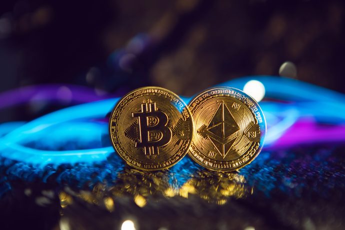 Zwei goldfarbene Münzen mit den Logos von Bitcoin und Ethereum stehen aufrecht auf einer strukturierten Oberfläche, mit verschwommenen blauen und violetten Lichtern im Hintergrund, die die Bedeutung von Bitcoin in der Welt der digitalen Währungen unterstreichen.