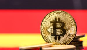 Ein goldfarbener physischer Bitcoin steht aufrecht zwischen anderen Münzen, mit einem verschwommenen roten, gelben und schwarzen Hintergrund, der an Deutschland erinnert.
