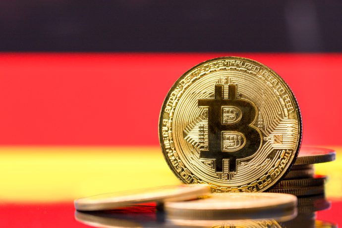 Ein goldfarbener physischer Bitcoin steht aufrecht zwischen anderen Münzen, mit einem verschwommenen roten, gelben und schwarzen Hintergrund, der an Deutschland erinnert.