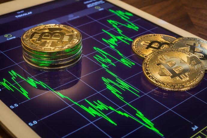 Stapel von physischen Bitcoin-Token liegen auf einem Tablet, das ein schwankendes Kryptowährungspreisdiagramm mit grünen Bitcoin-Grafiklinien anzeigt.