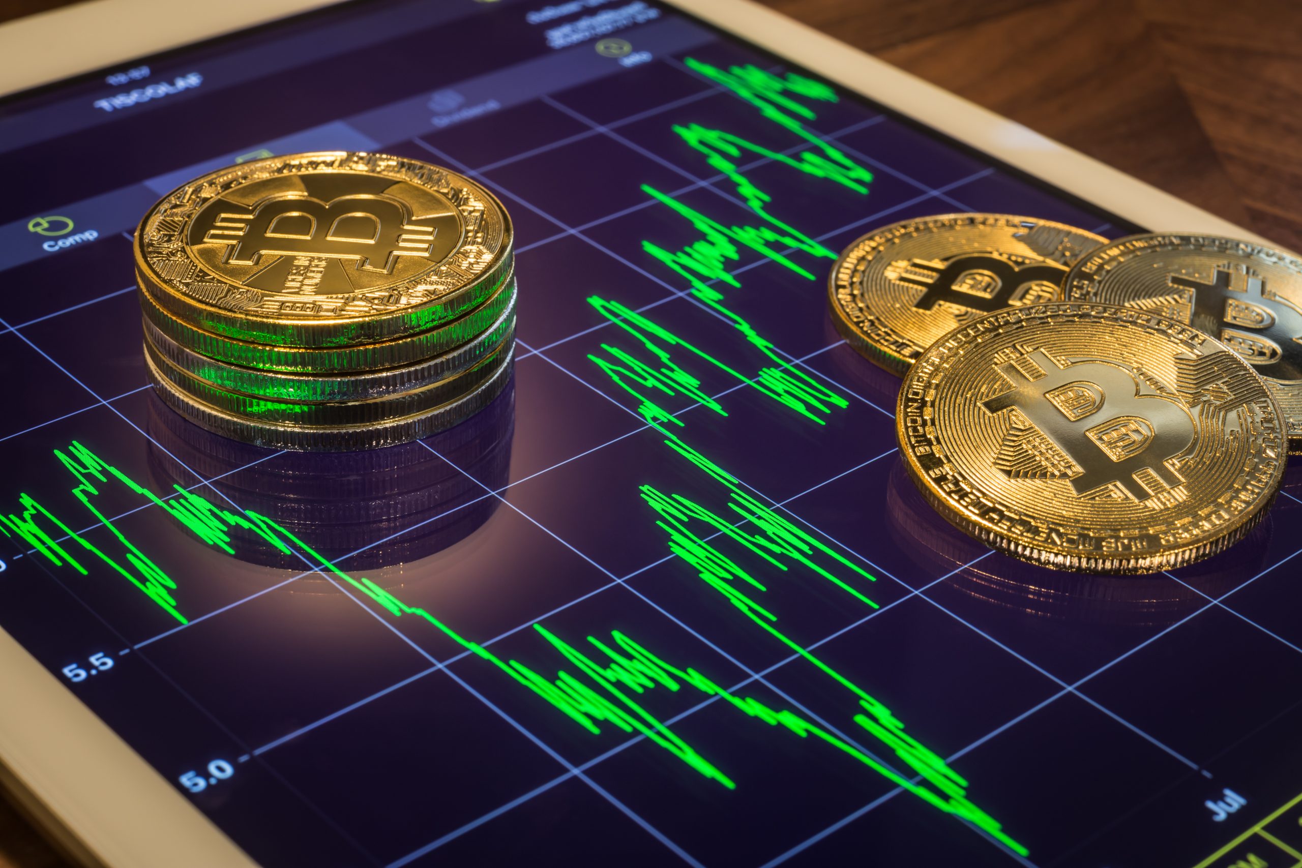 Stapel von physischen Bitcoin-Token liegen auf einem Tablet, das ein schwankendes Kryptowährungspreisdiagramm mit grünen Bitcoin-Grafiklinien anzeigt.