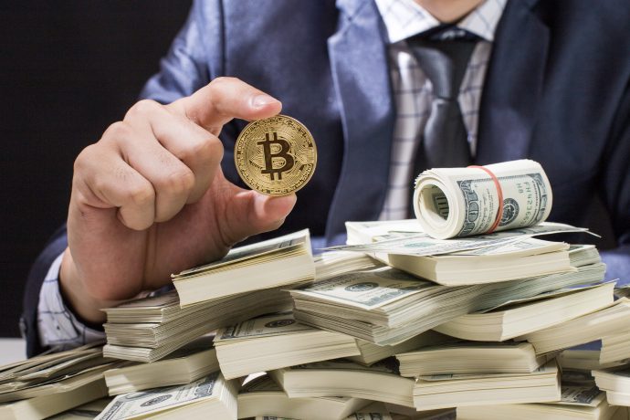 Eine Person im Anzug hält einen Bitcoin-Token über einem großen Stapel von US-Dollar-Scheinen, auf dem eine Rolle Bargeld liegt, was den Kontrast zwischen Kryptowährung und herkömmlicher Währung verdeutlicht.