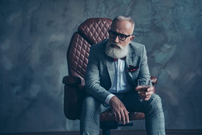 Ein älterer Mann mit weißem Bart und Brille sitzt in einem braunen Ledersessel, trägt einen grau karierten Anzug, hält ein Glas Whiskey in der Hand und denkt über seine Bitcoin-Investitionen nach.