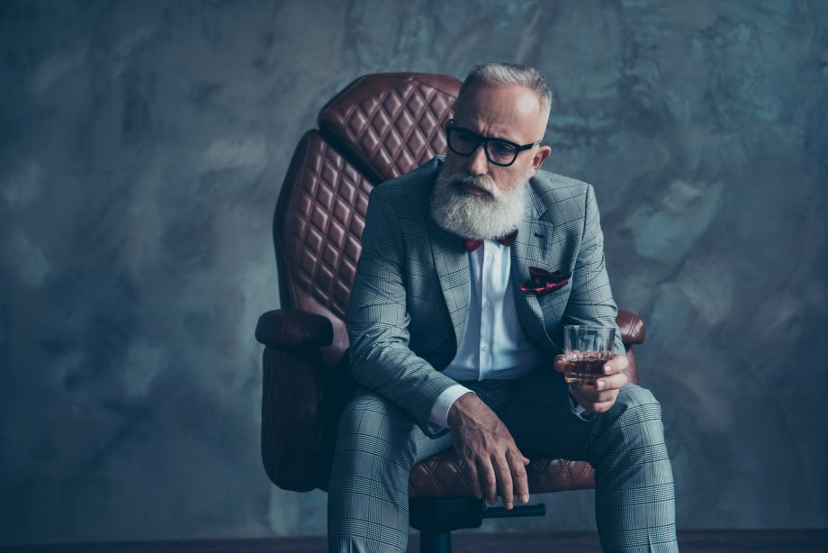 Ein älterer Mann mit weißem Bart und Brille sitzt in einem braunen Ledersessel, trägt einen grau karierten Anzug, hält ein Glas Whiskey in der Hand und denkt über seine Bitcoin-Investitionen nach.