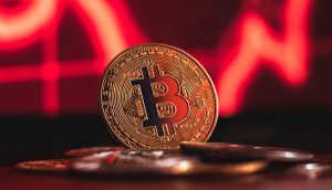 Eine physische Darstellung einer Bitcoin-Münze steht auf einem Haufen Münzen, mit einem roten, abwärts gerichteten Graphen im unscharfen Hintergrund, der die jüngsten Trends des Krypto-Marktupdates hervorhebt.