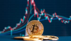 Ein Stapel Bitcoin-Münzen liegt vor einem Bildschirm, der ein Finanzdiagramm mit roten und blauen Linien anzeigt und die volatile Kursbewegung von Bitcoin verdeutlicht.