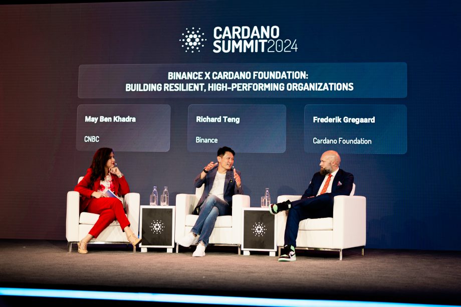Drei Diskussionsteilnehmer sitzen auf der Bühne des Cardano Summit 2025 in Berlin, hinter ihnen ist ein Bildschirm mit ihren Namen und Zugehörigkeiten zu sehen: CNBC, Binance und Cardano Foundation.