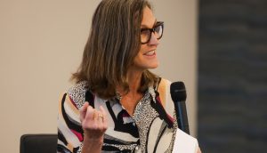 Eine Frau mit Brille spricht in ein Handmikrofon über Krypto, während sie Papiere in der Hand hält und mit ihrer linken Hand in einer modernen Innenumgebung gestikuliert.