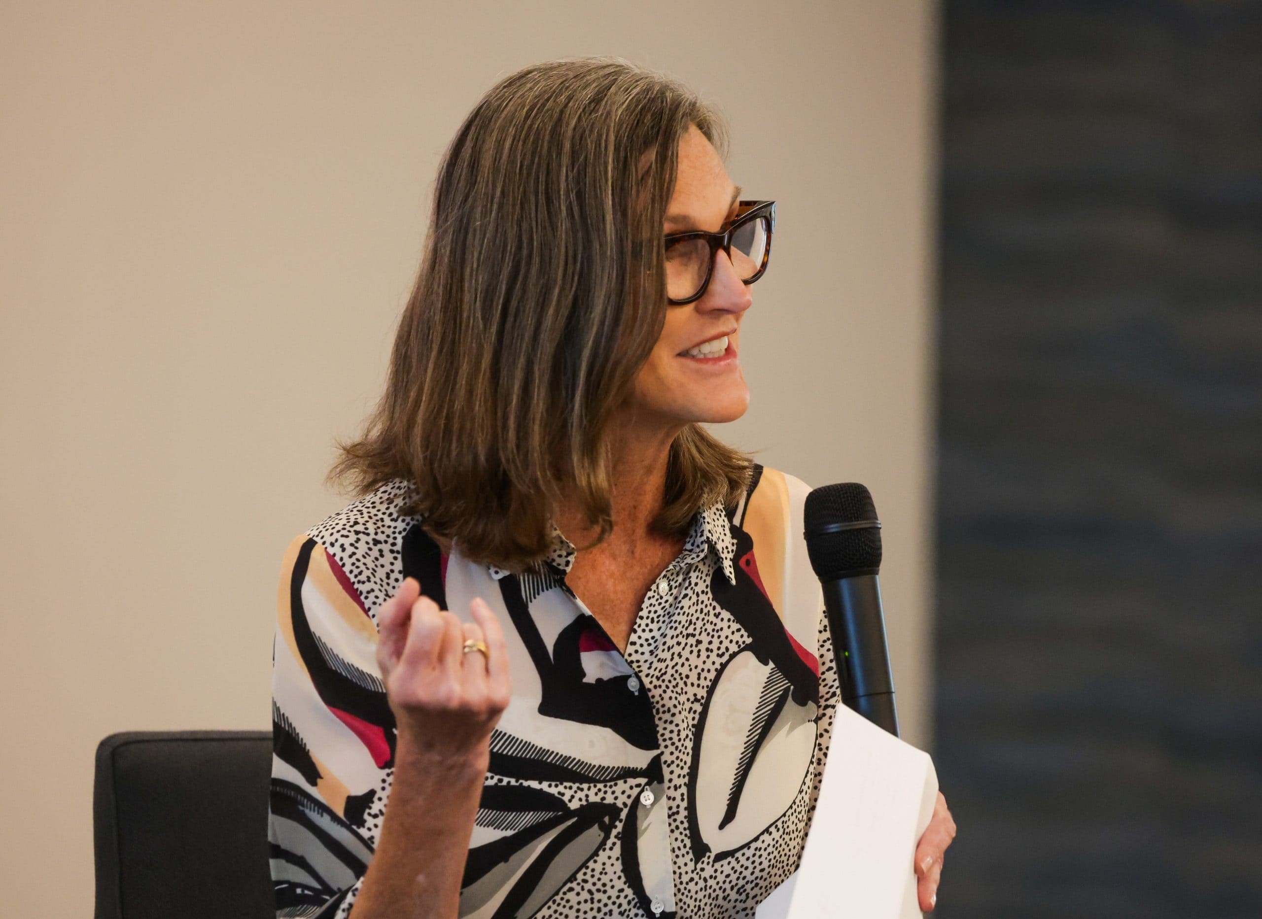 Eine Frau mit Brille spricht in ein Handmikrofon über Krypto, während sie Papiere in der Hand hält und mit ihrer linken Hand in einer modernen Innenumgebung gestikuliert.