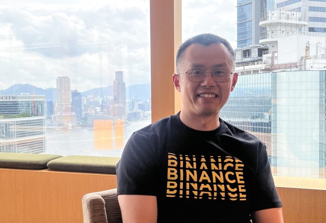 CZ steht lächelnd vor einem Fenster und trägt stolz ein Binance-T-Shirt.