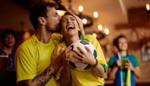 Ein Mann küsst eine lachende Frau auf die Stirn, während sie einen Fußball in der Hand hält; beide tragen gelbe Trikots in einer belebten Halle und feiern ihre Lieblingsmannschaft und die wachsende Begeisterung für SportFi.