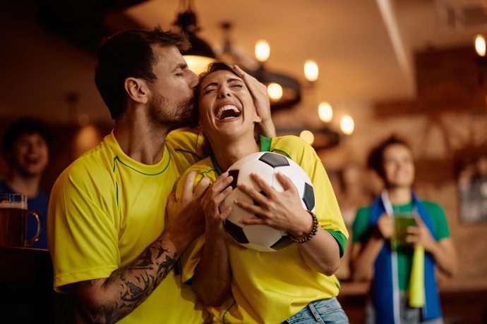 Ein Mann küsst eine lachende Frau auf die Stirn, während sie einen Fußball in der Hand hält; beide tragen gelbe Trikots in einer belebten Halle und feiern ihre Lieblingsmannschaft und die wachsende Begeisterung für SportFi.