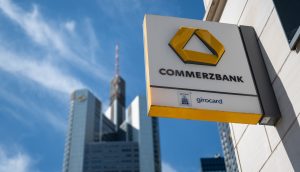 Commerzbank-Zeichen auf einem Gebäude vor dem Hintergrund eines modernen Wolkenkratzers und eines klaren blauen Himmels.